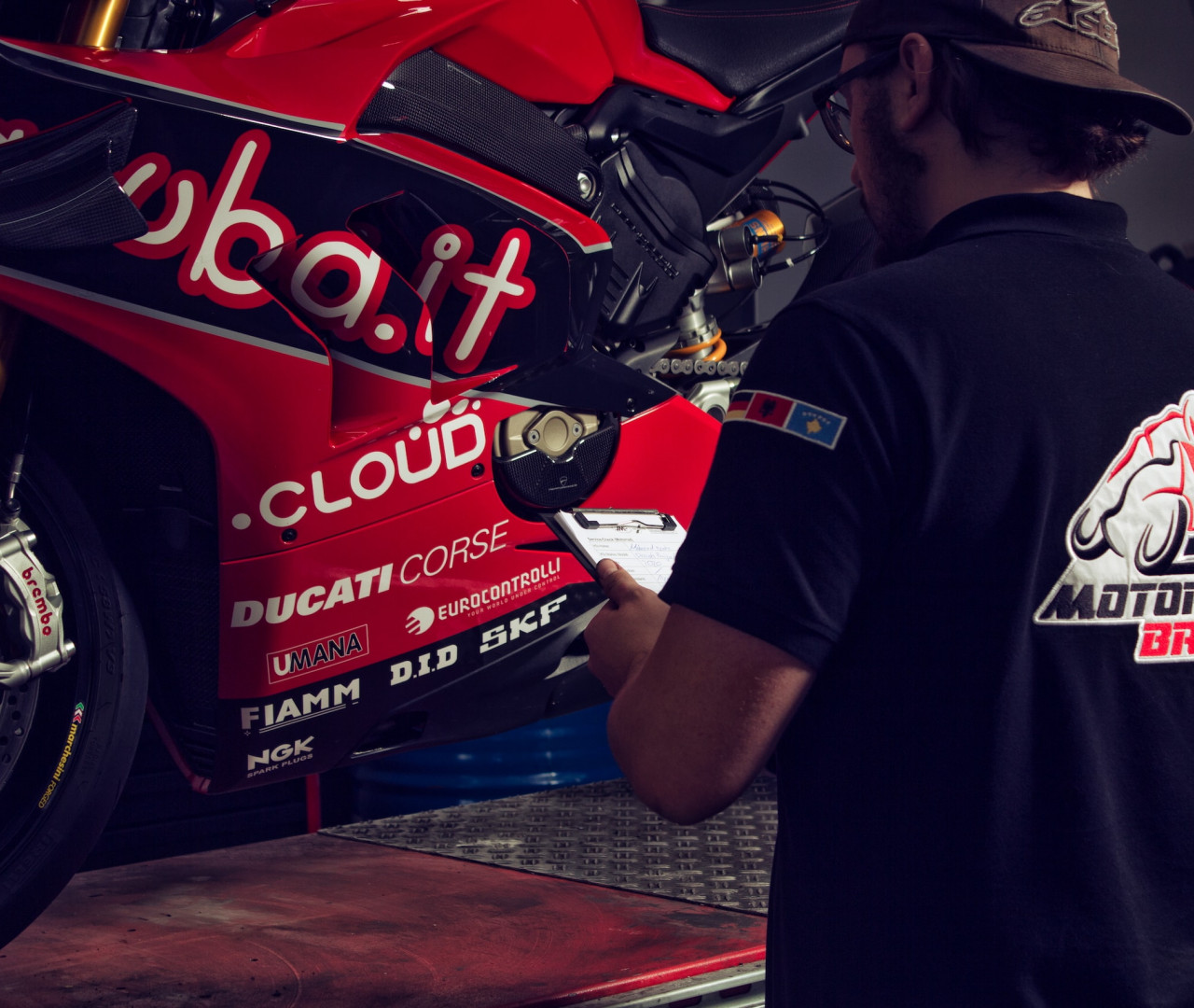 Ein Mechaniker in schwarzem T-Shirt mit dem Motorrad Braha Logo auf dem Rücken arbeitet an einem roten Ducati Rennmotorrad. Das Motorrad trägt prominente Sponsorenaufkleber, darunter 'sbai.it', 'CLOUD' und 'DUCATI CORSE', sowie weitere Markierungen von Sponsoren wie SKF, DID, FIAMM und NGK. Der Mechaniker scheint Notizen auf einem Klemmbrett zu machen, während er neben dem auf einem Werkstattboden stehenden Motorrad steht. Die goldene Öhlins-Federung ist am Vorderrad deutlich zu erkennen.